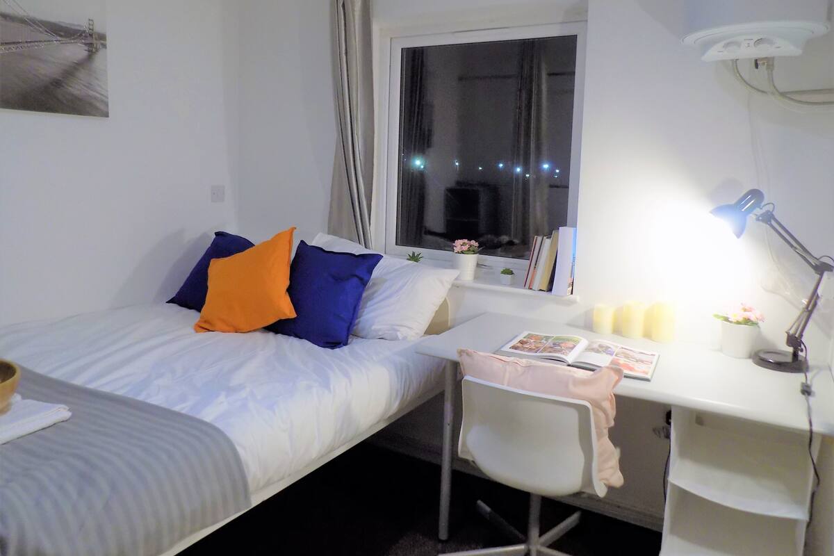 A bedroom with a bed, a desk, a chair, a window, and a nightstand with a lamp.
