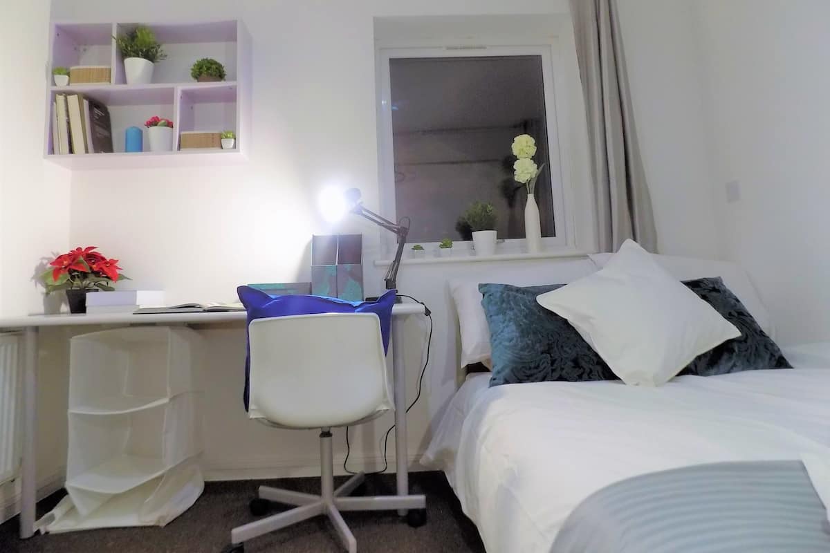 A bedroom with a bed, a desk with a chair, a window with curtains, and a shelf with plants and books.