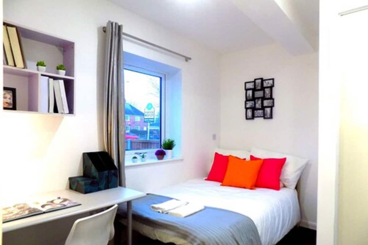 A small, well-lit room with a bed, a desk, and a chair. There is a window with a view of buildings outside.