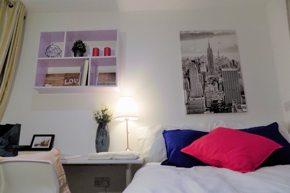 A hotel room with a bed, a desk with a lamp, a chair, a shelf with decorative items, and a cityscape picture on the wall.