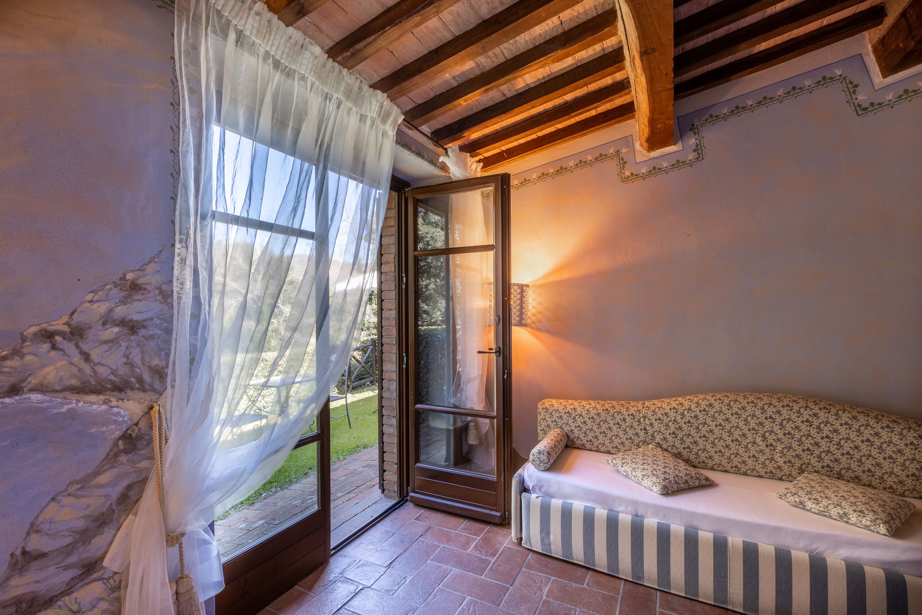 Une pièce chaleureuse avec un plafond en bois, un mur en pierre, une porte vitrée donnant sur l’extérieur et un canapé agrémenté de coussins à motifs floraux.