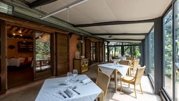 Uma área de refeições interna com mesas para dois, janelas amplas e vista para o exterior.