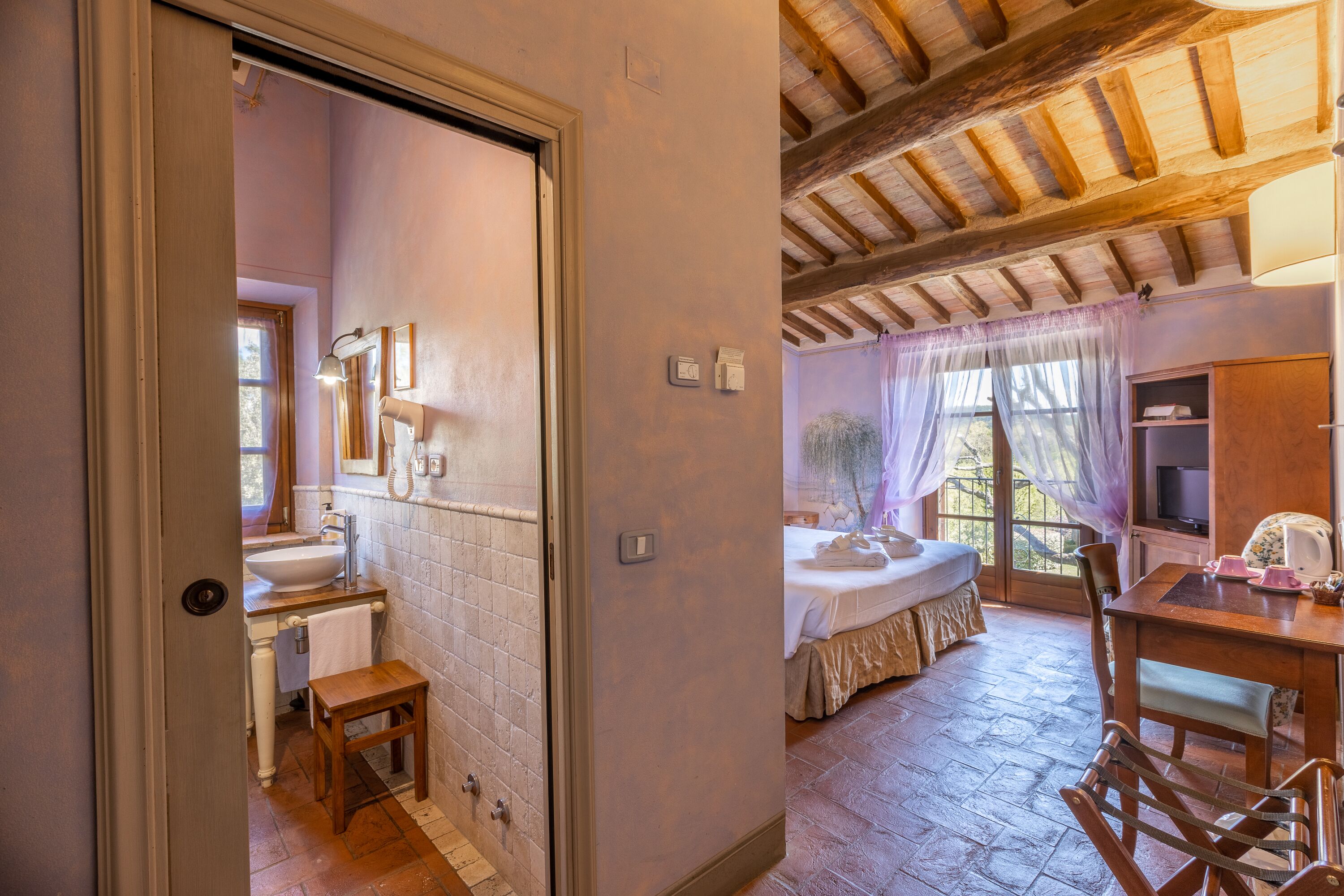 Una habitación con cama, una mesa de madera, televisor y baño con lavamanos y espejo.