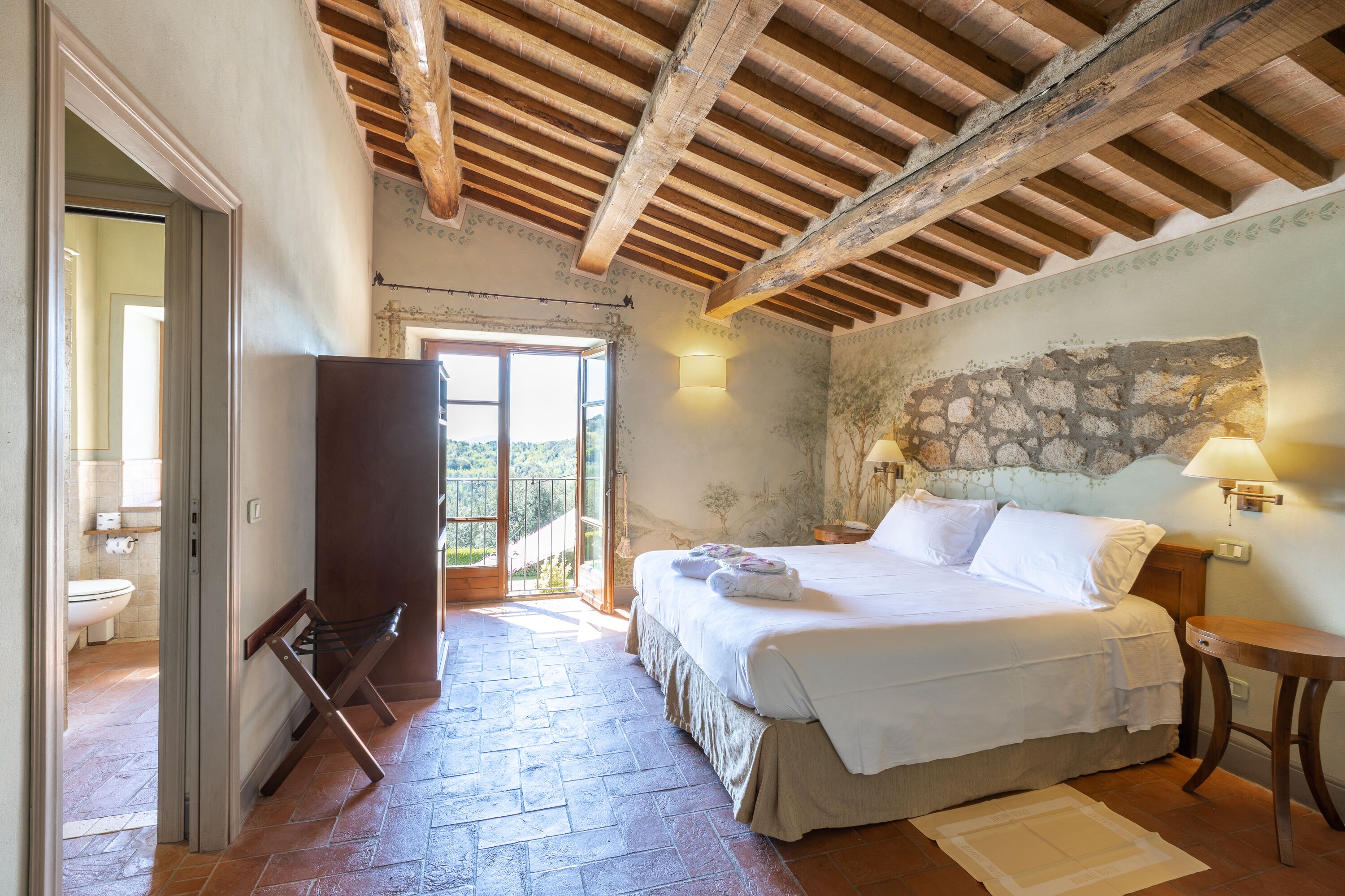 Un dormitorio con cabecera de piedra, cama con ropa blanca, banca de madera y vista a través de un ventanal.