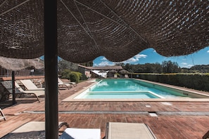 Una piscina con sillones de descanso y sombrillas, rodeada de pavimento de ladrillo y un seto.