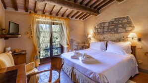 Une chambre avec un mur en pierre, un lit recouvert de draps blancs, une chaise en bois, un bureau avec un ordinateur et une fenêtre avec des rideaux.