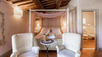 A bedroom with a four-poster bed, two white armchairs, a small table with a bottle and glasses, and a bathroom visible through an open door.