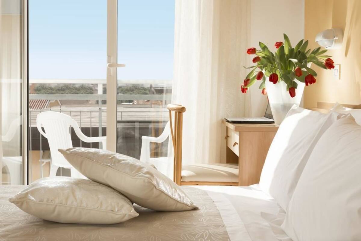 A hotel room with a bed, white pillows, a nightstand with a vase of red tulips, a laptop, and a view of the outdoors through large windows.