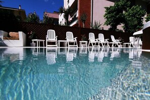 A pool with clear water, surrounded by white lounge chairs and a wicker sofa. - Hotel Bellerofonte (Rimini)