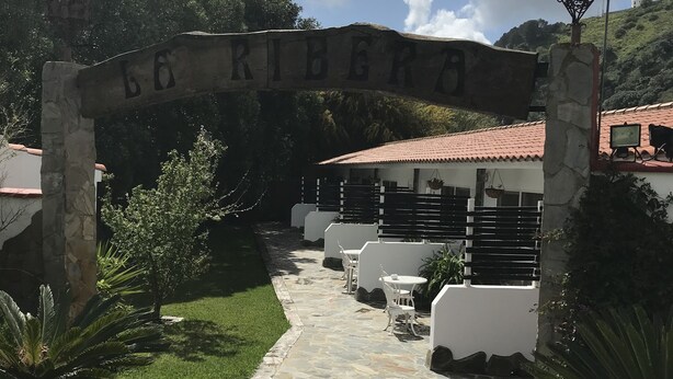 En stenlagd gång leder fram till en vit byggnad med ett tak täckt av tegelpannor. Byggnaden omges av grönska och en stenvalvbåge med namnet "RIBEIRA" ovanför.