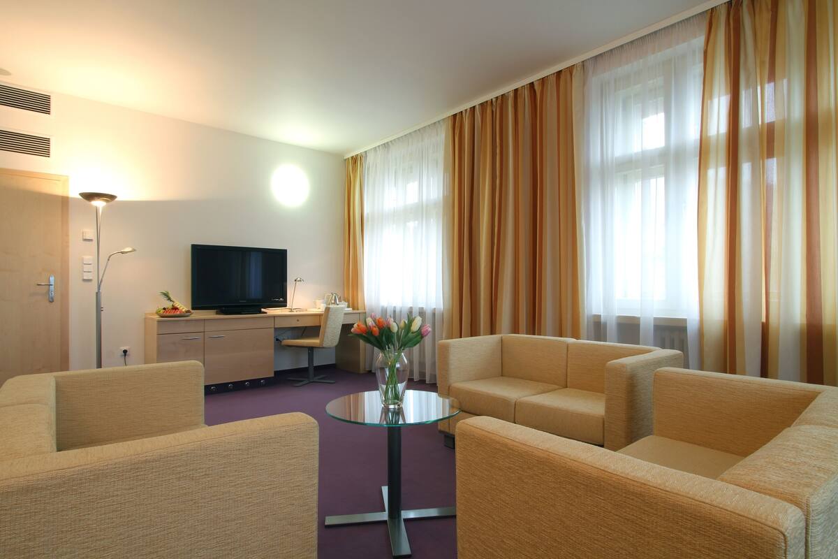 Habitación de hotel con un conjunto de sofás, una mesita redonda con un jarrón de flores, un televisor en un escritorio y cortinas grandes.