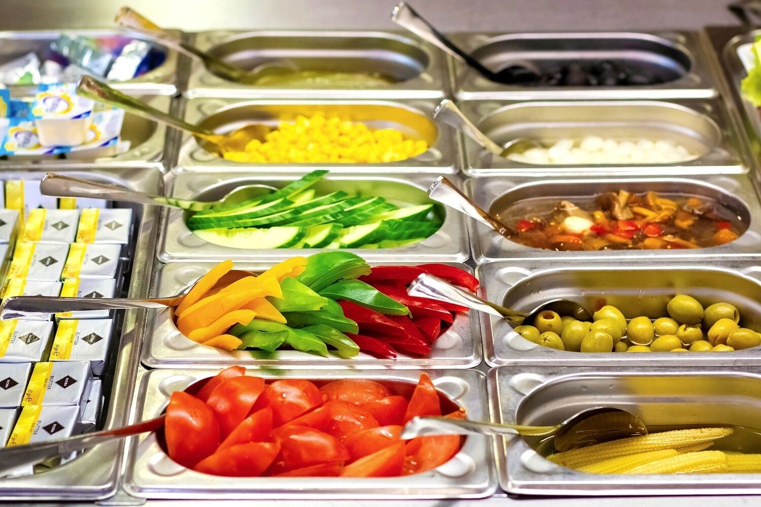 Un bufé con diferentes alimentos, como maíz, pepinos, pimientos, aceitunas y tomates.