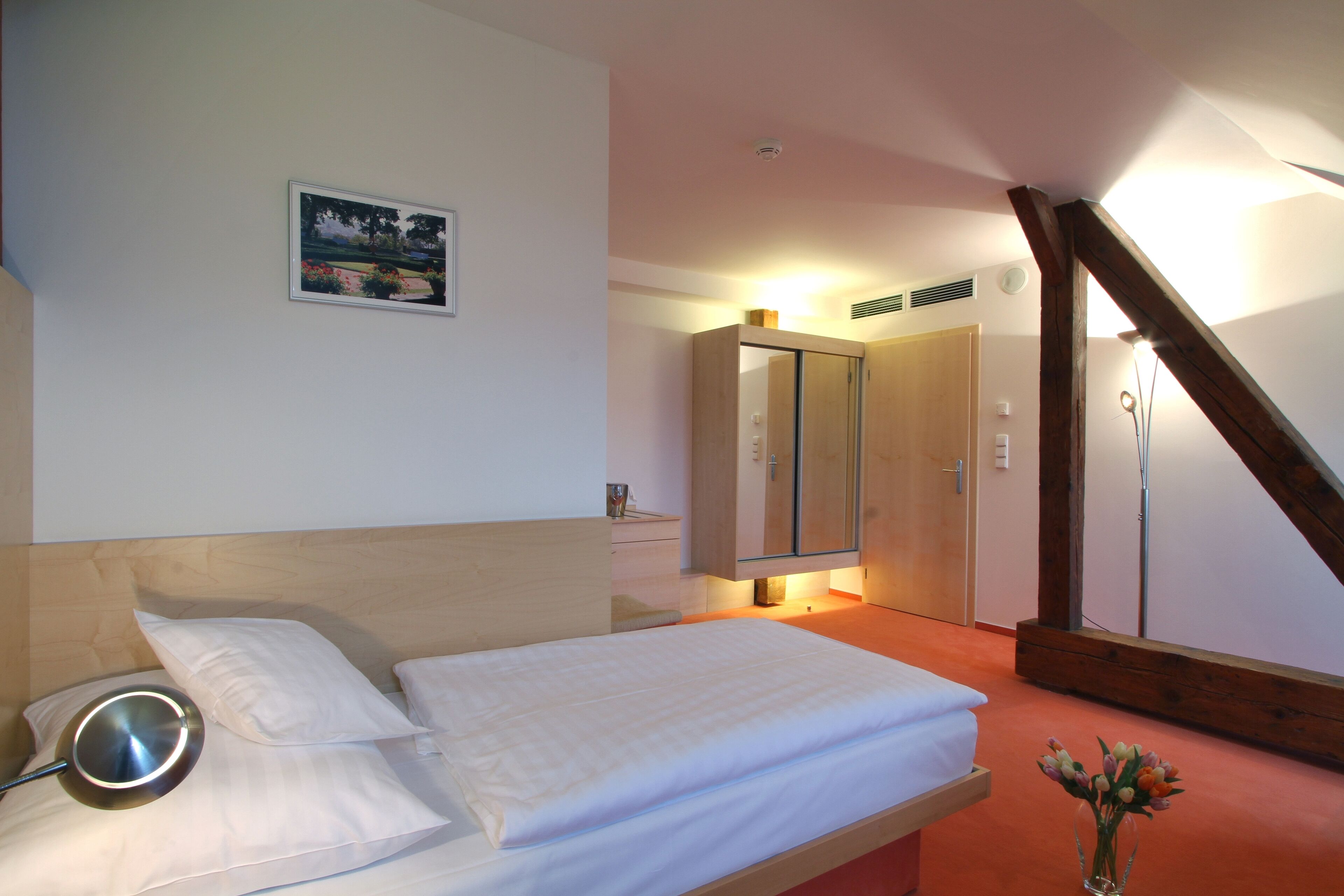 A bedroom with a bed, a bedside table, a mirror, a picture on the wall, and a vase with flowers.