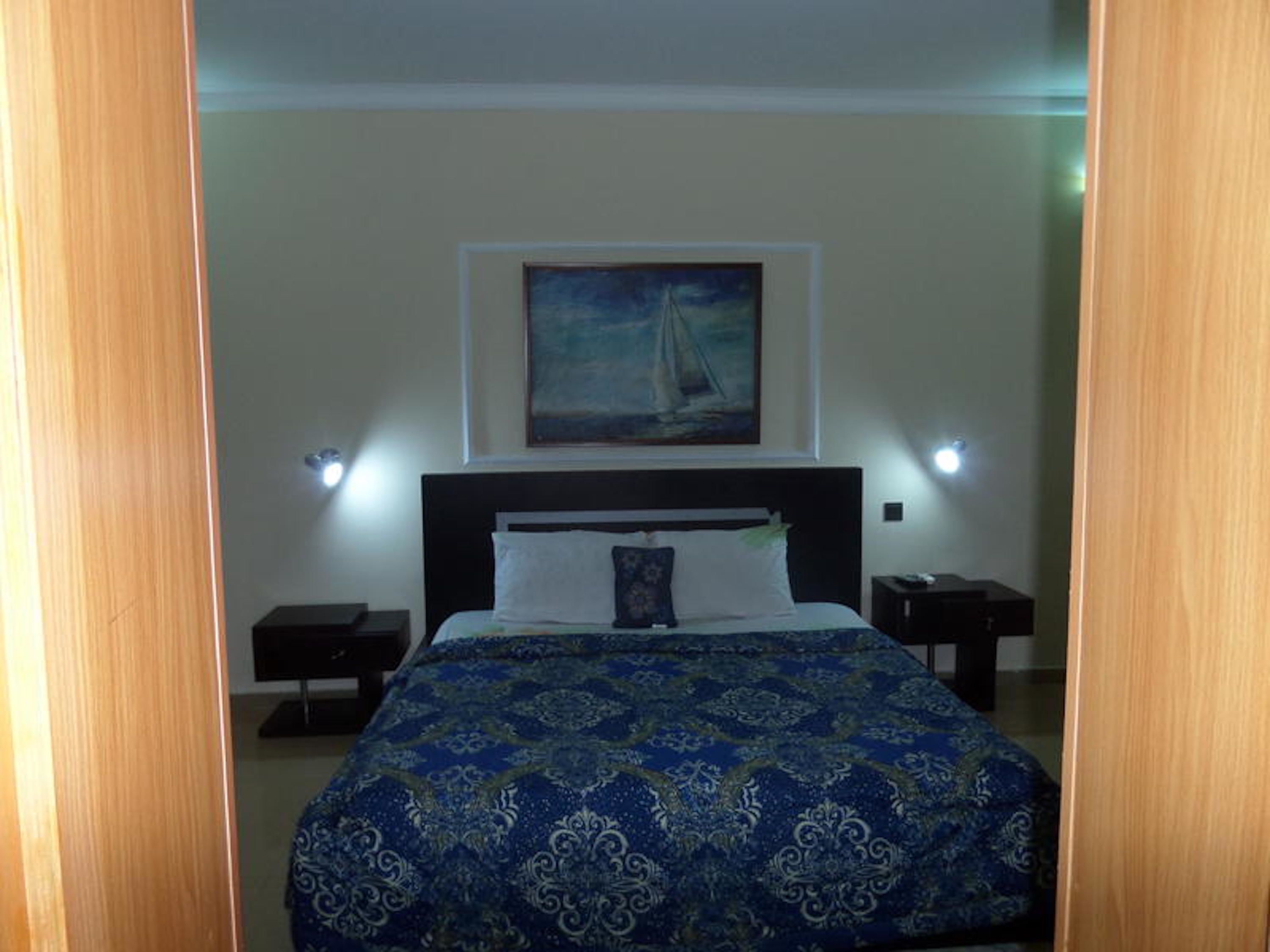 A bedroom with a bed, a painting of a sailboat on the wall, and two bedside tables with lamps.