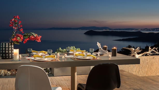 A table set for dinner with candles, plates, and a vase of flowers, overlooking a calm sea and distant hills. - Marathon Blue (Marathon)
