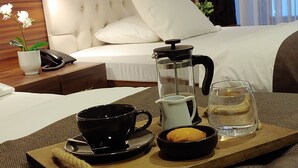 Ein ordentlich angerichteter Frühstückswagen mit Kaffeekanne, Tasse und Schüssel auf einer Holzbrettchen, platziert auf einer strukturierten Matte in einem Hotelzimmer mit Backsteinwand, einem gesteppten Kopfteil und einem Bett mit weißen Leinen.
