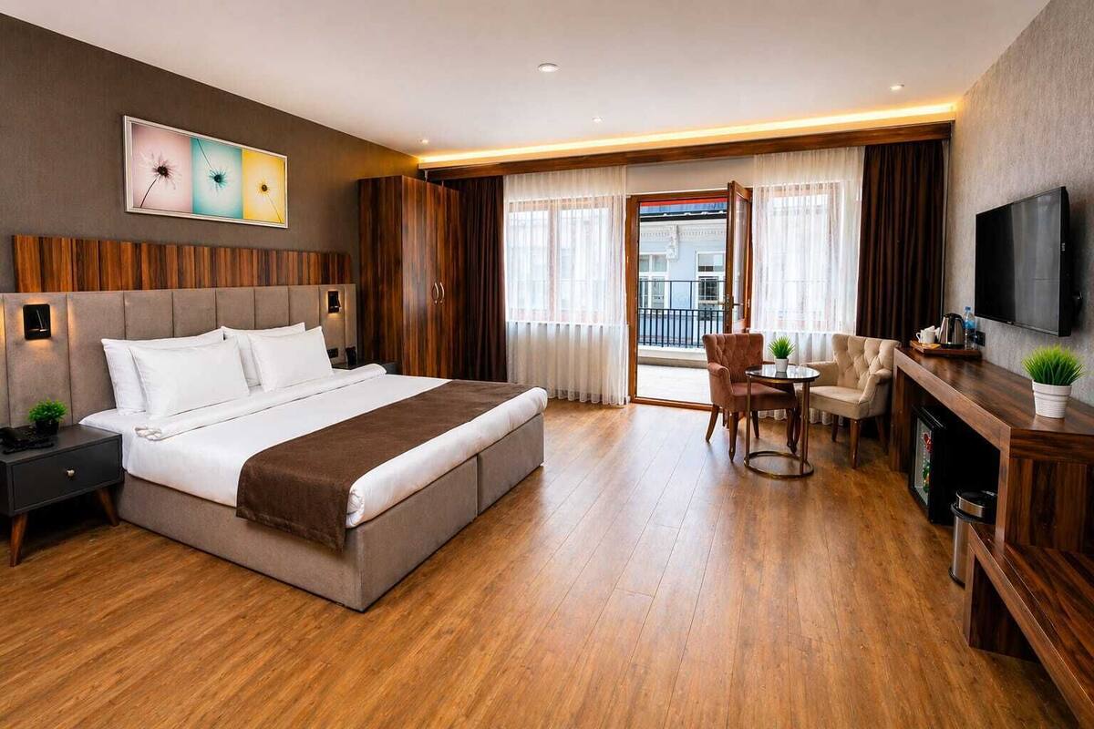 A hotel room with a large bed, a desk with a chair, a TV, and a balcony with curtains.