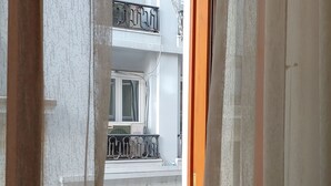 Blick durch ein Fenster mit Vorhang, der einen Balkon mit Geländer, einen Blumentopf und einen Tisch mit Teekanne, Tassen und Krug zeigt.