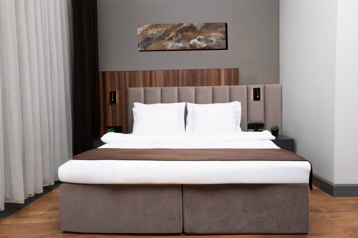 A modern hotel room with a large bed, a wooden headboard, two bedside tables, and a painting on the wall.