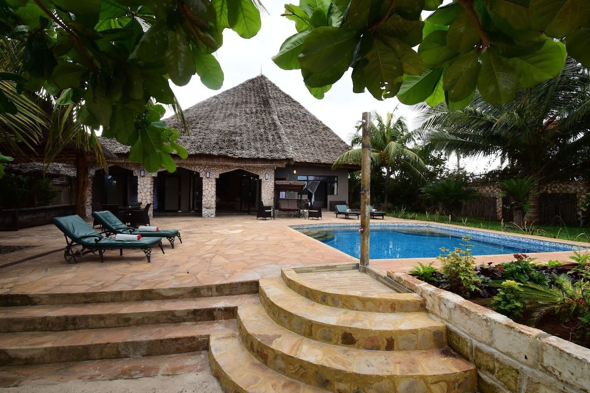 Un área de piscina con sillones de descanso, una construcción con techo de paja y una escalera de piedra que conduce a la piscina.