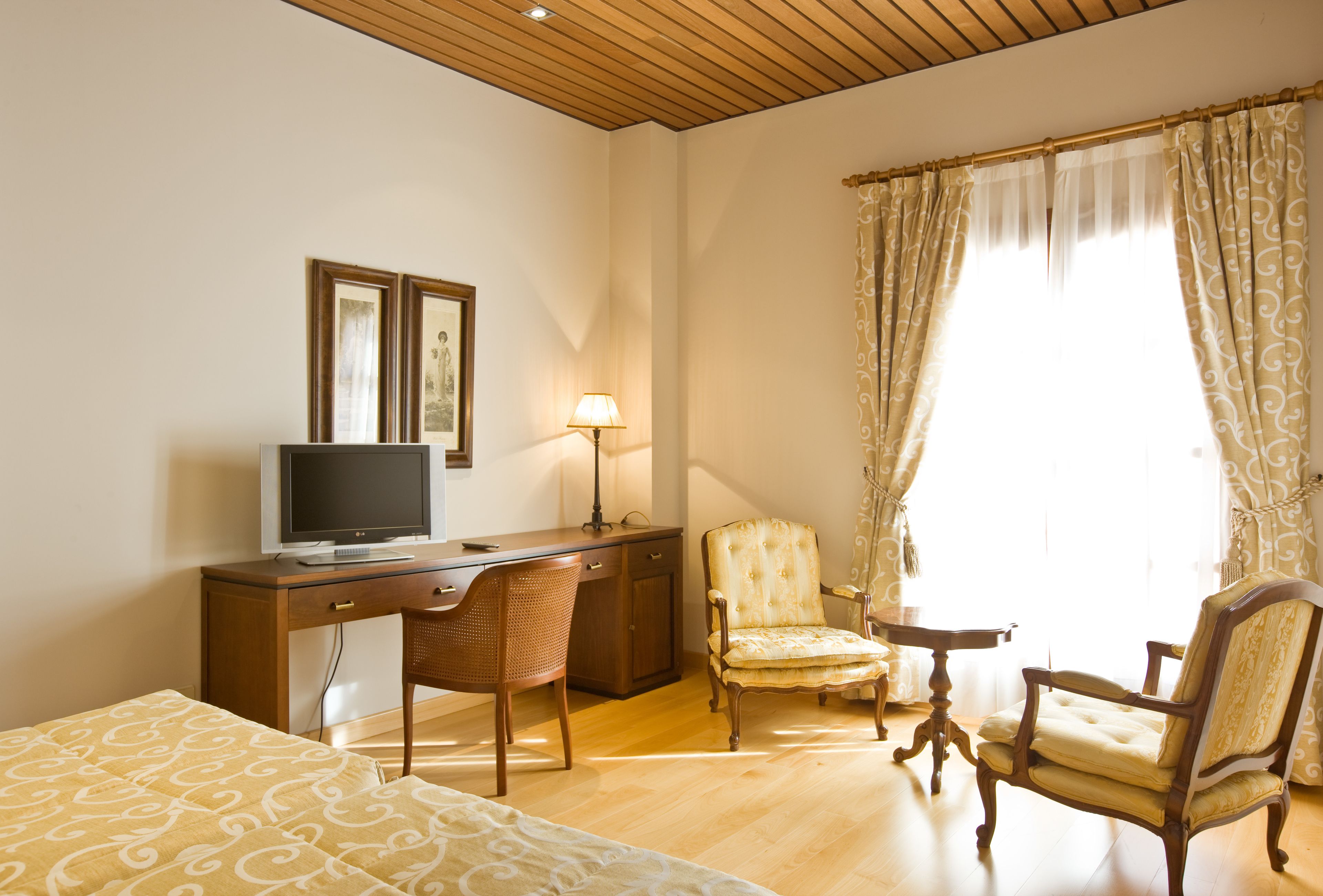 Una habitación de hotel con un escritorio de madera, televisión, lámpara, dos sillas y una cama con ropa de cama estampada.