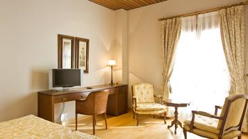 Una habitación de hotel con un escritorio de madera, televisión, lámpara, dos sillas y una cama con ropa de cama estampada.
