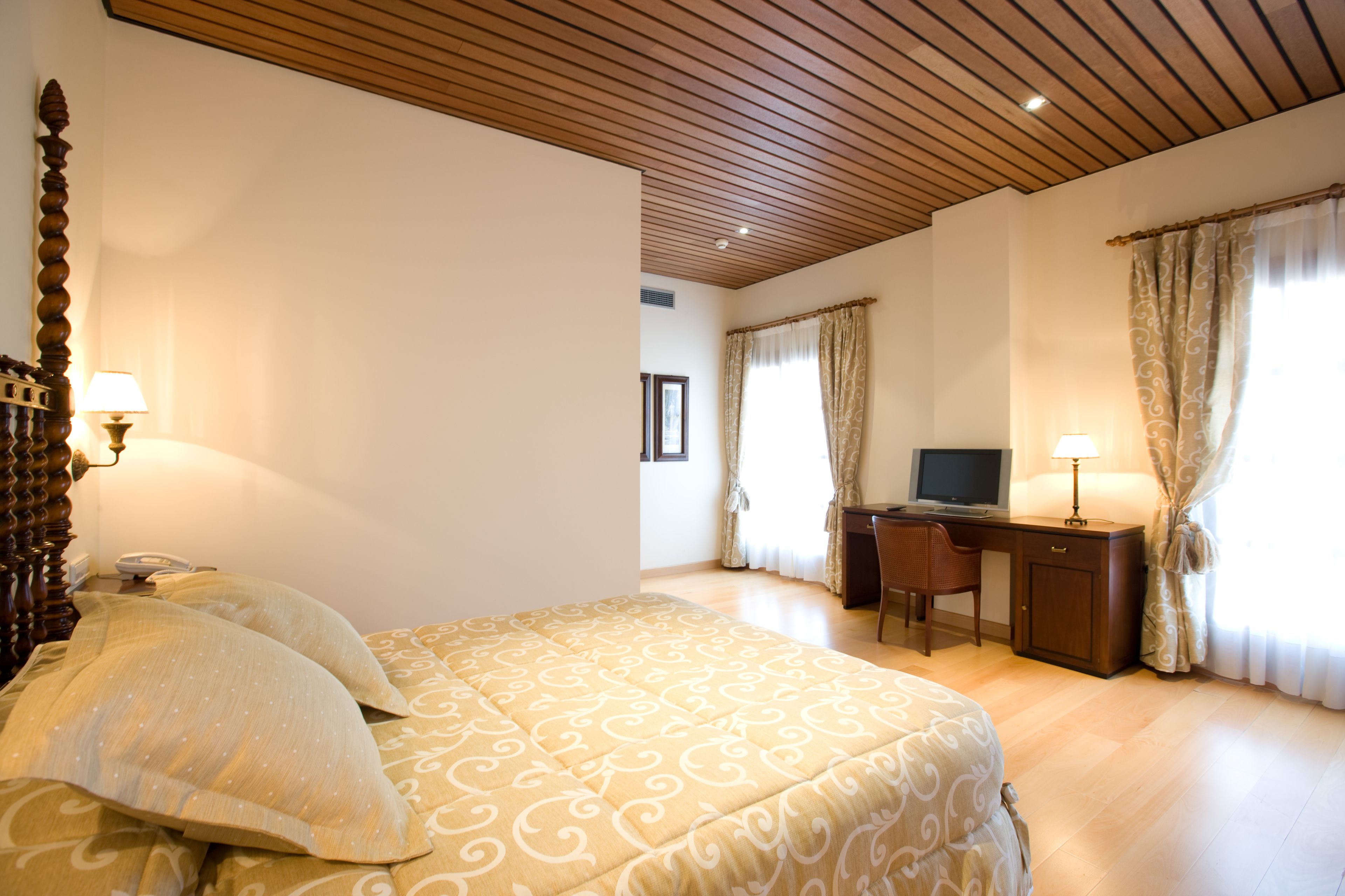 Habitación de hotel con techo de madera, cama con ropa de cama estampada, escritorio con computadora y cortinas en las ventanas.