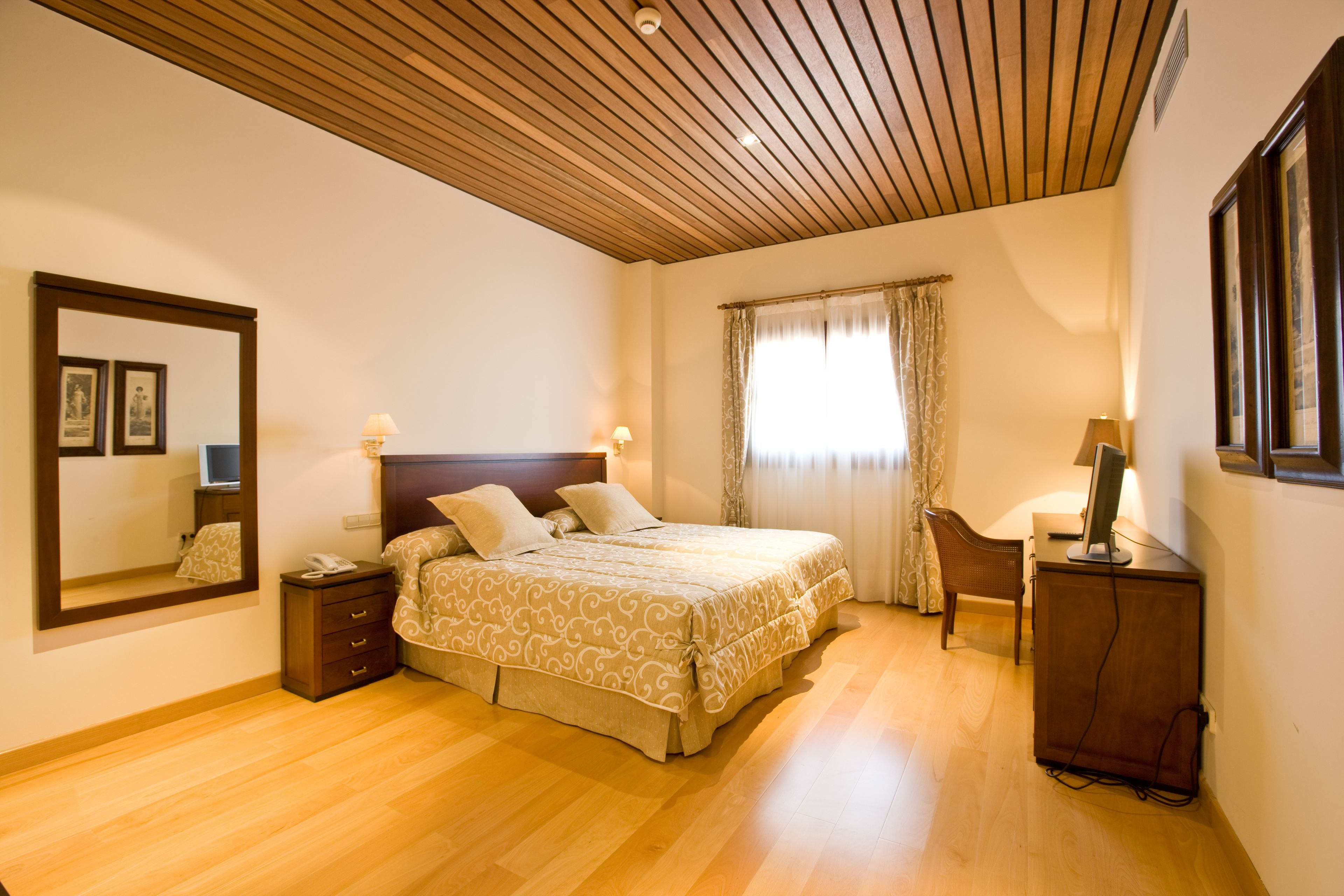 Un dormitorio con techo de madera, una cama con colcha estampada, una mesita de noche con lámpara, una silla y un escritorio con computadora.