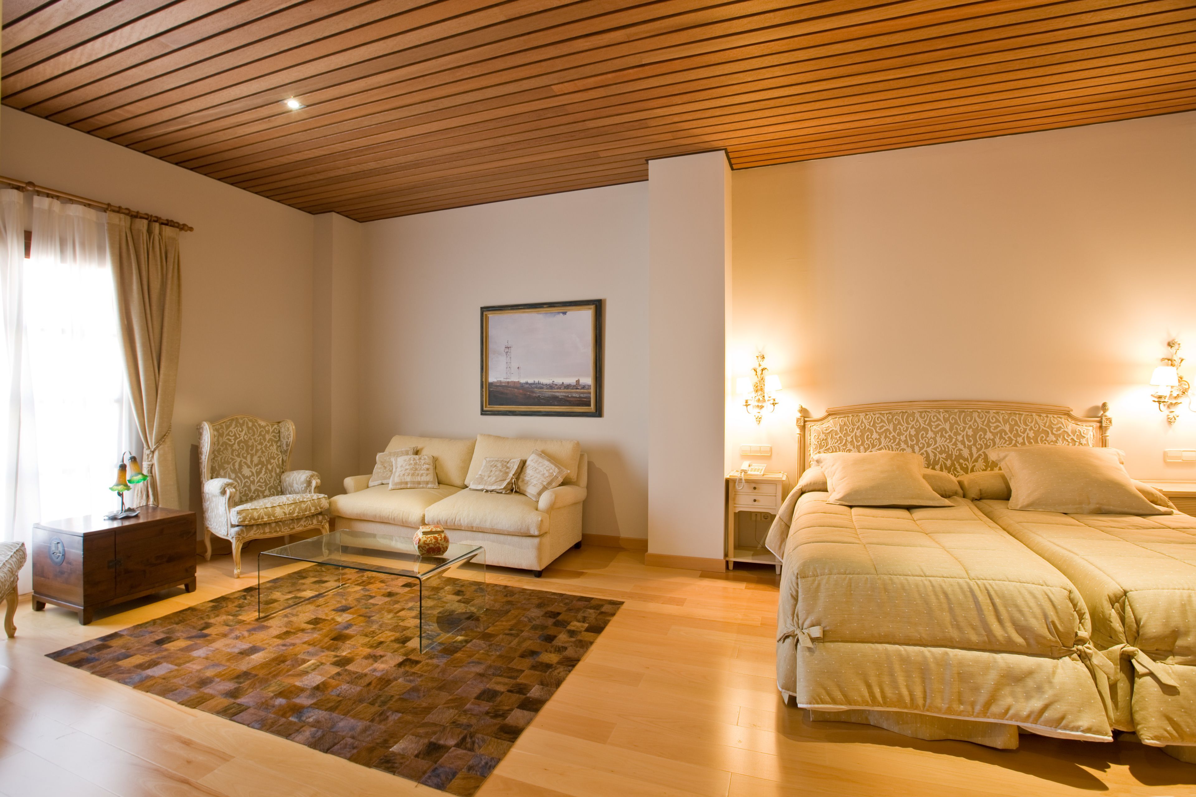Een slaapkamer met een houten plafond, een bed met hoofdeinde, een bank, een bijzettafeltje en een vloerkleed.