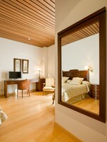 A hotel room with a wooden ceiling, a large bed with a wooden headboard, a desk with a computer, a chair, and a bedside table with a lamp.