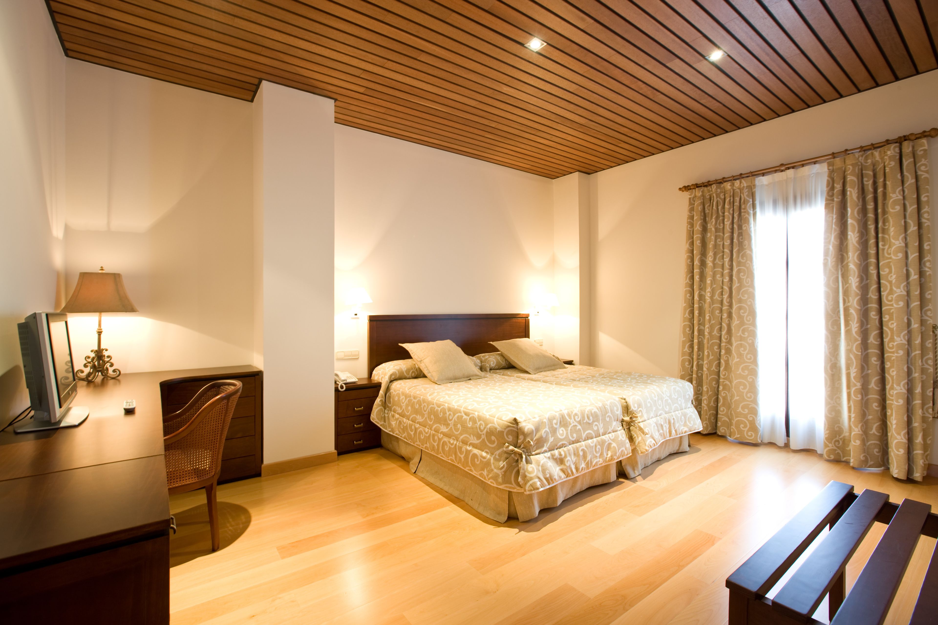 Un dormitorio con techo de madera, cama con ropa de cama estampada, escritorio con lámpara y una silla.