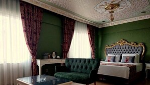 Una habitación con pared verde, cama con cabecera dorada, un sillón verde, una mesa con cubierta de vidrio y un escritorio blanco.