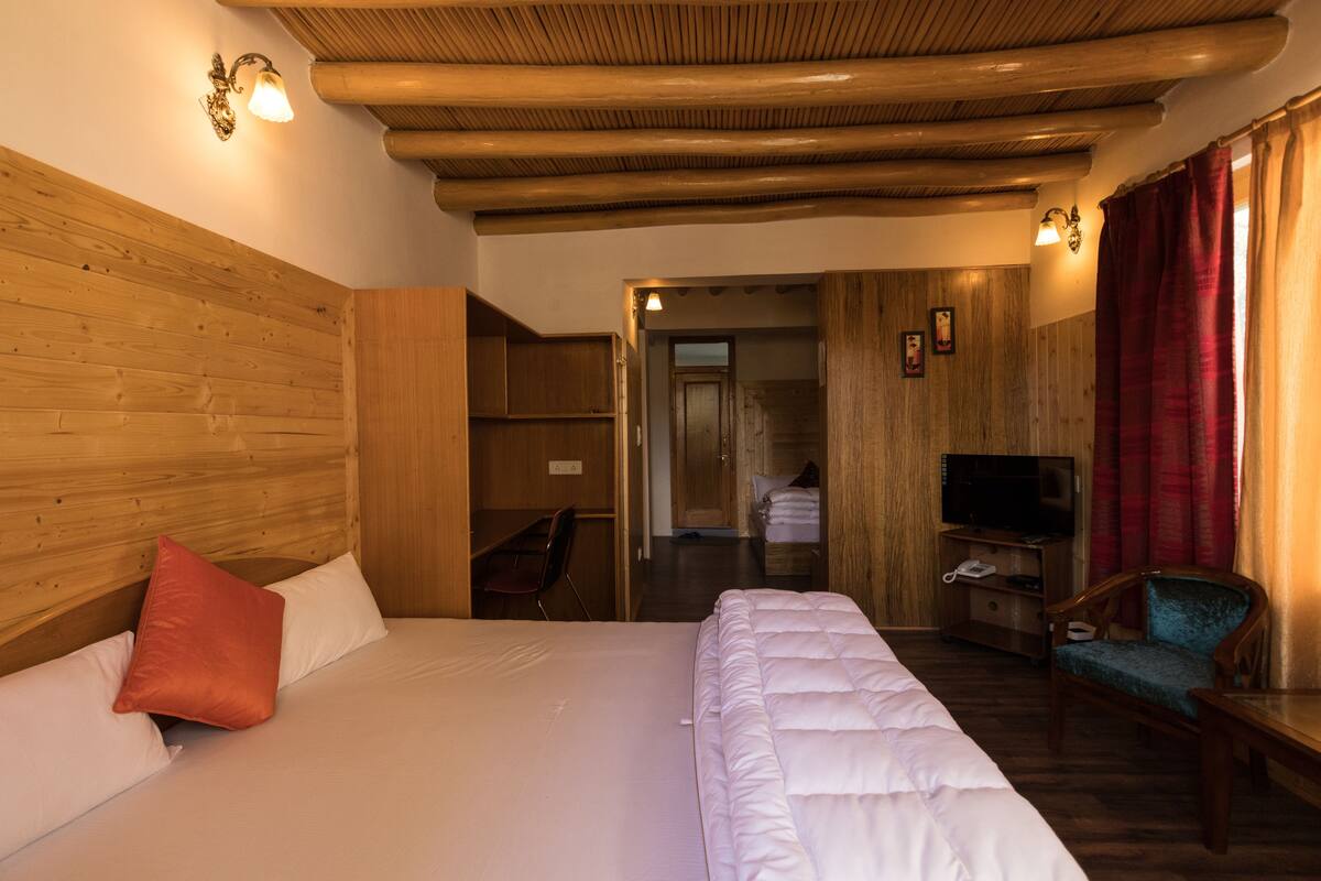 A hotel room with a wooden headboard, a bed with white bedding, a red pillow, a wooden TV stand with a television, and a chair.