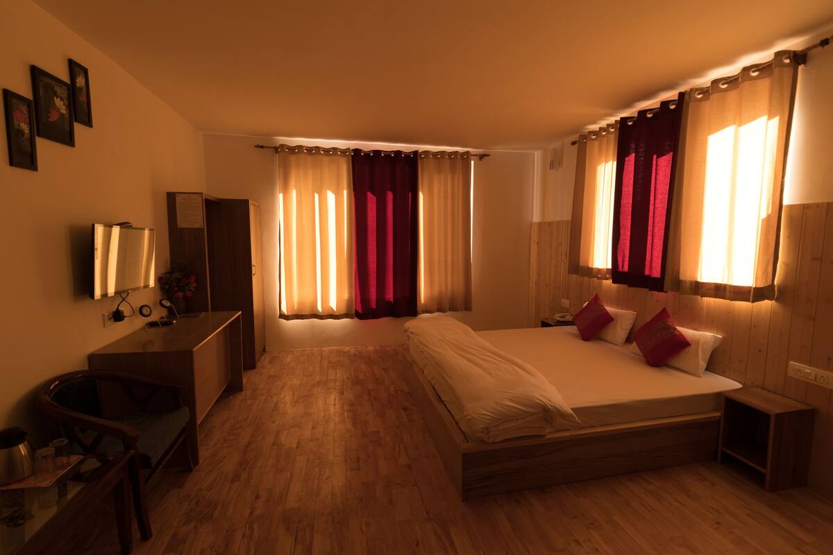 A hotel room with a wooden floor, a bed with white bedding and red pillows, a desk with a mirror, and a window with curtains.