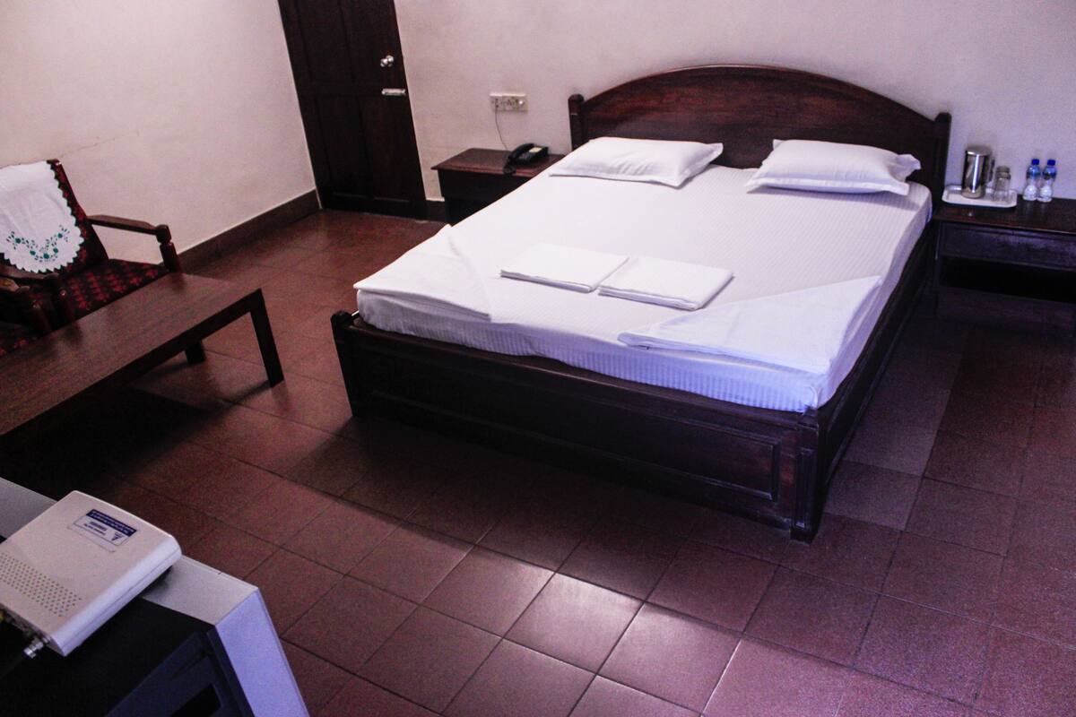 A neatly made bed with pillows and folded towels, a bedside table with a phone and a water bottle, and a chair with a decorative cloth.