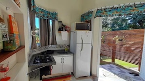 A compact kitchen with a gas stove, white refrigerator, and a microwave. There is a small window with curtains, and a wooden fence outside. - Bangalô Red Moon (Florianópolis)
