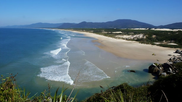 A coastal landscape with a sandy beach, clear blue waters, and distant mountains. - Bangalô Red Moon (Florianópolis)