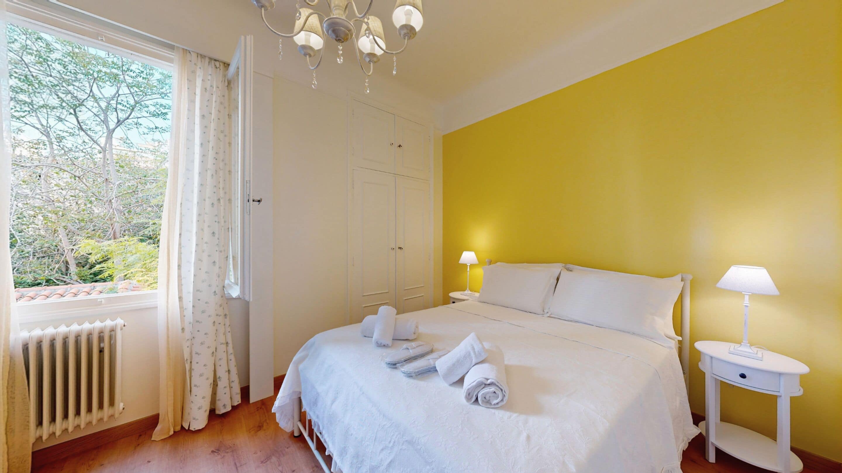 Un dormitorio con una cama, dos mesitas de noche con lámparas, una ventana con cortinas y un radiador.