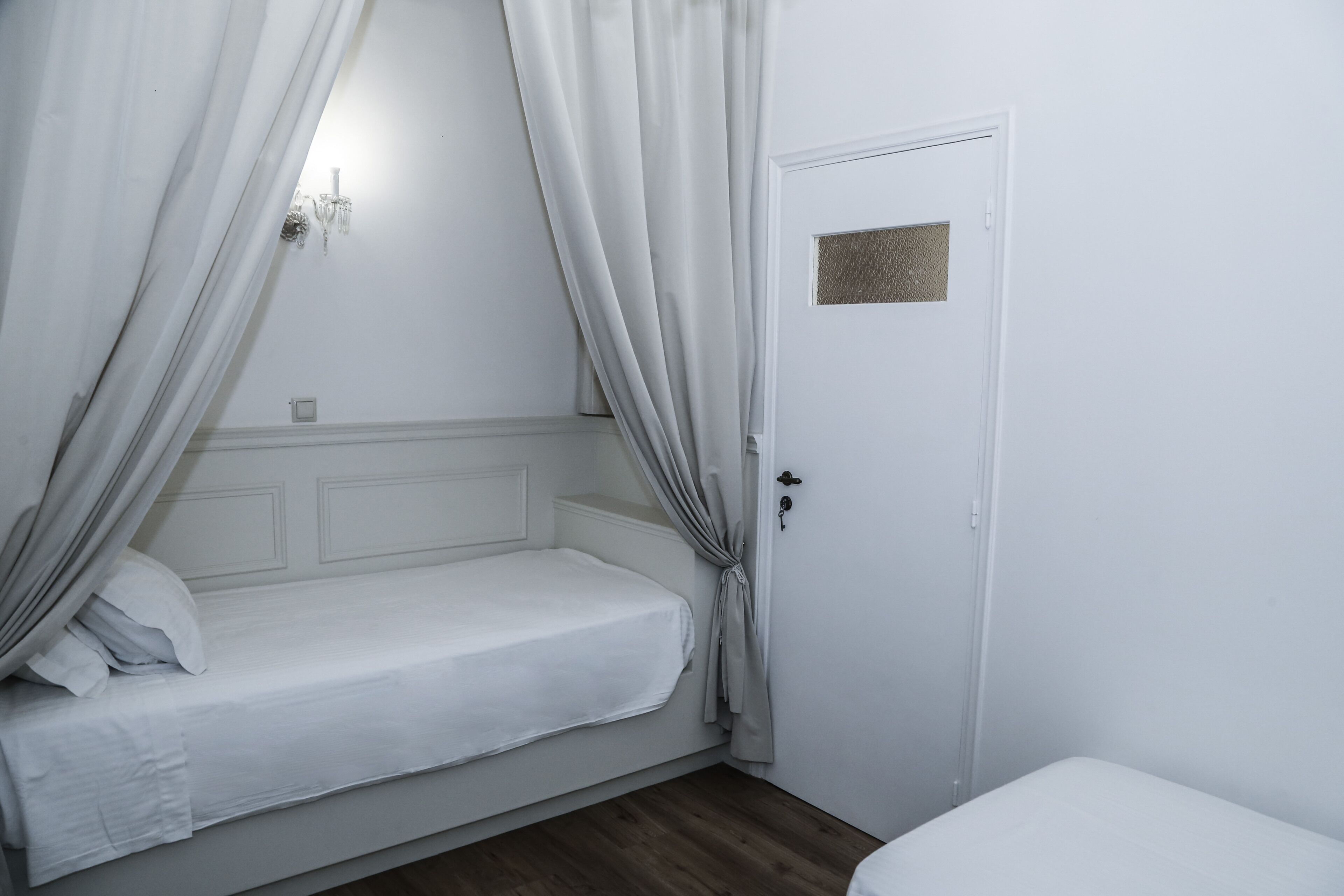 Une petite chambre avec un lit simple, une tête de lit blanche, une porte avec une petite fenêtre et un plancher en bois.