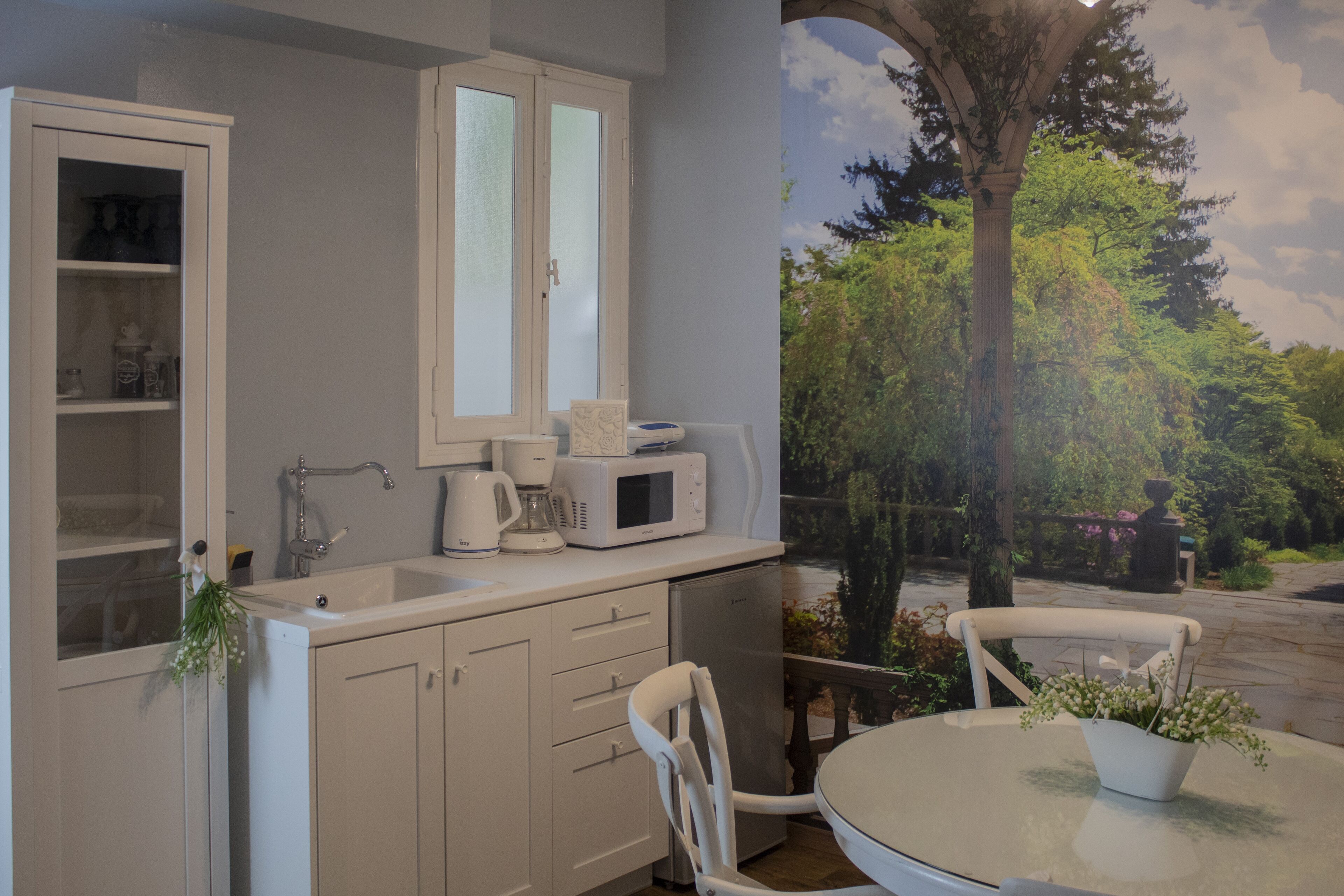 A kitchen with a white countertop, a sink, a microwave, and a coffee maker. There is a window with frosted glass and a view of trees and a stone pathway.