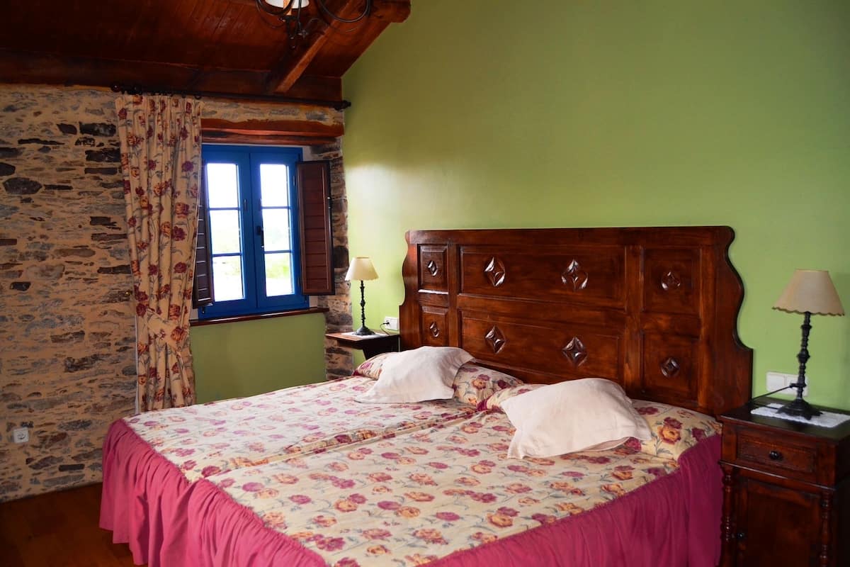 A bedroom with a wooden bed, floral bedspread, a stone wall, a window with blue shutters, and a green wall.