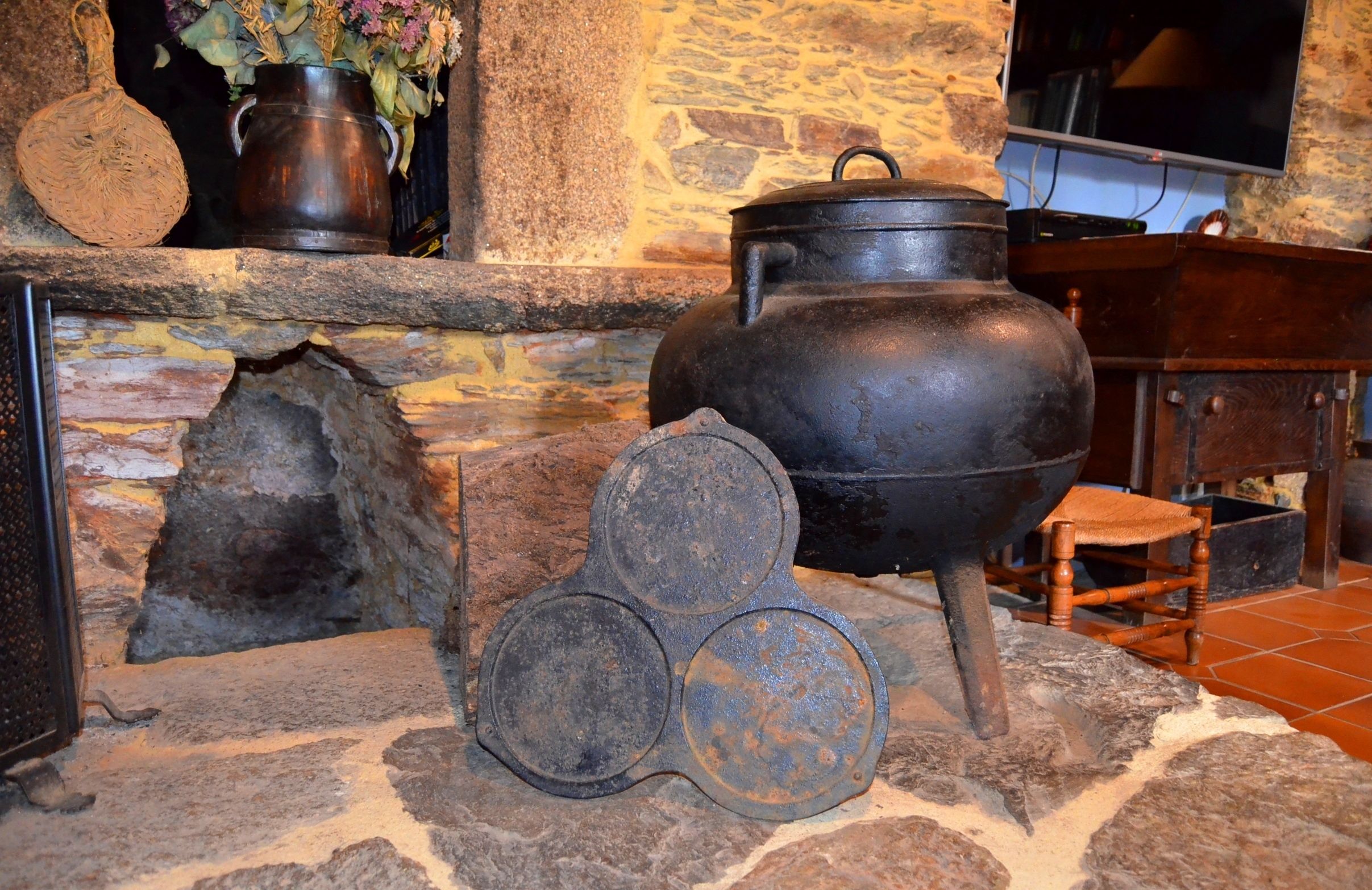 A rustic stone fireplace with a large pot, a wooden stool, and a metal tray.