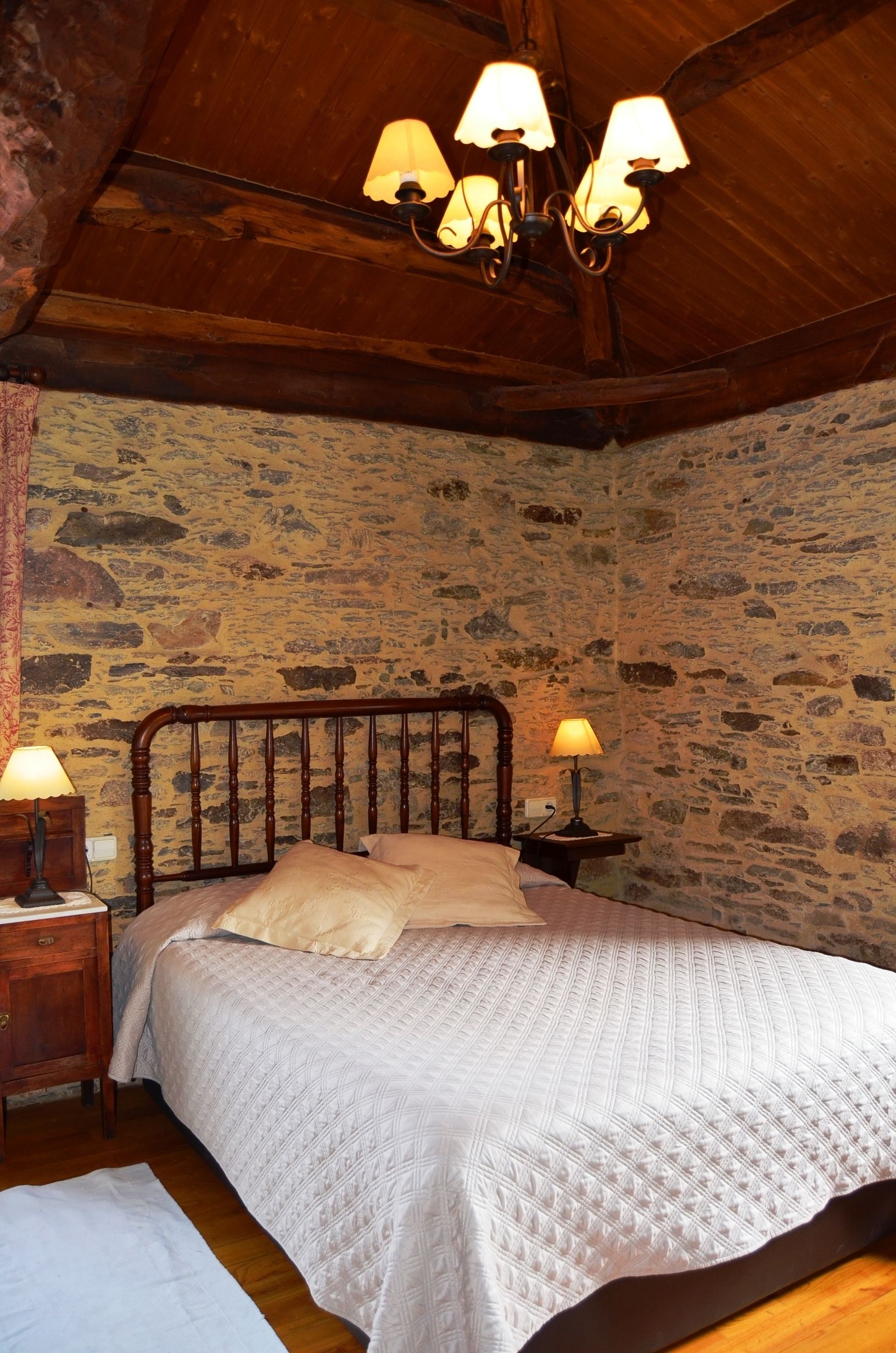 Een slaapkamer met een stenen muur, houten balken in het plafond, een bed met een gewatteerde witte dekbedovertrek en twee bedlampen.