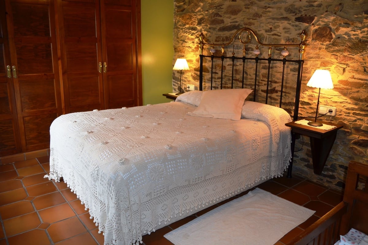 A neatly made bed with a white blanket and pillows, a wooden headboard, two bedside lamps, and a stone wall behind the bed.