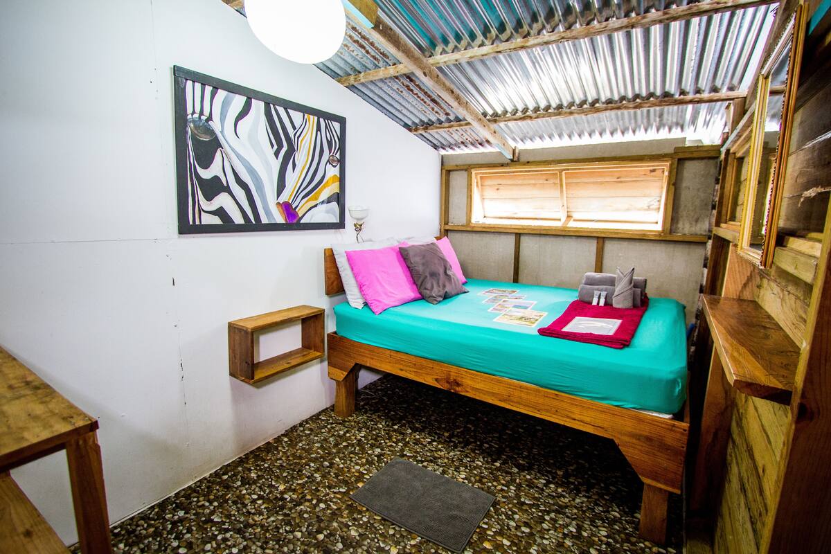 A small, rustic room with a wooden bed, a colorful abstract painting, a small wooden table, and a window with wooden shutters.