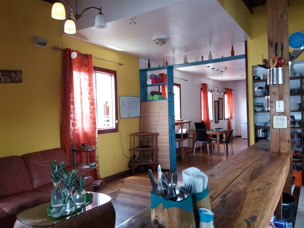 A cozy, well-lit room with a wooden dining table, chairs, and a sofa. There is a kitchen area with a refrigerator and various kitchen utensils.