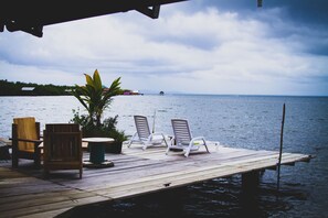A wooden dock with two lounge chairs and a table, overlooking a calm body of water with distant land and a cloudy sky.