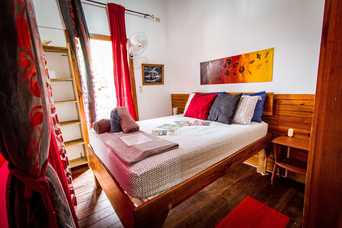 A bedroom with a wooden bed, red curtains, a fan, a painting, and a small table.