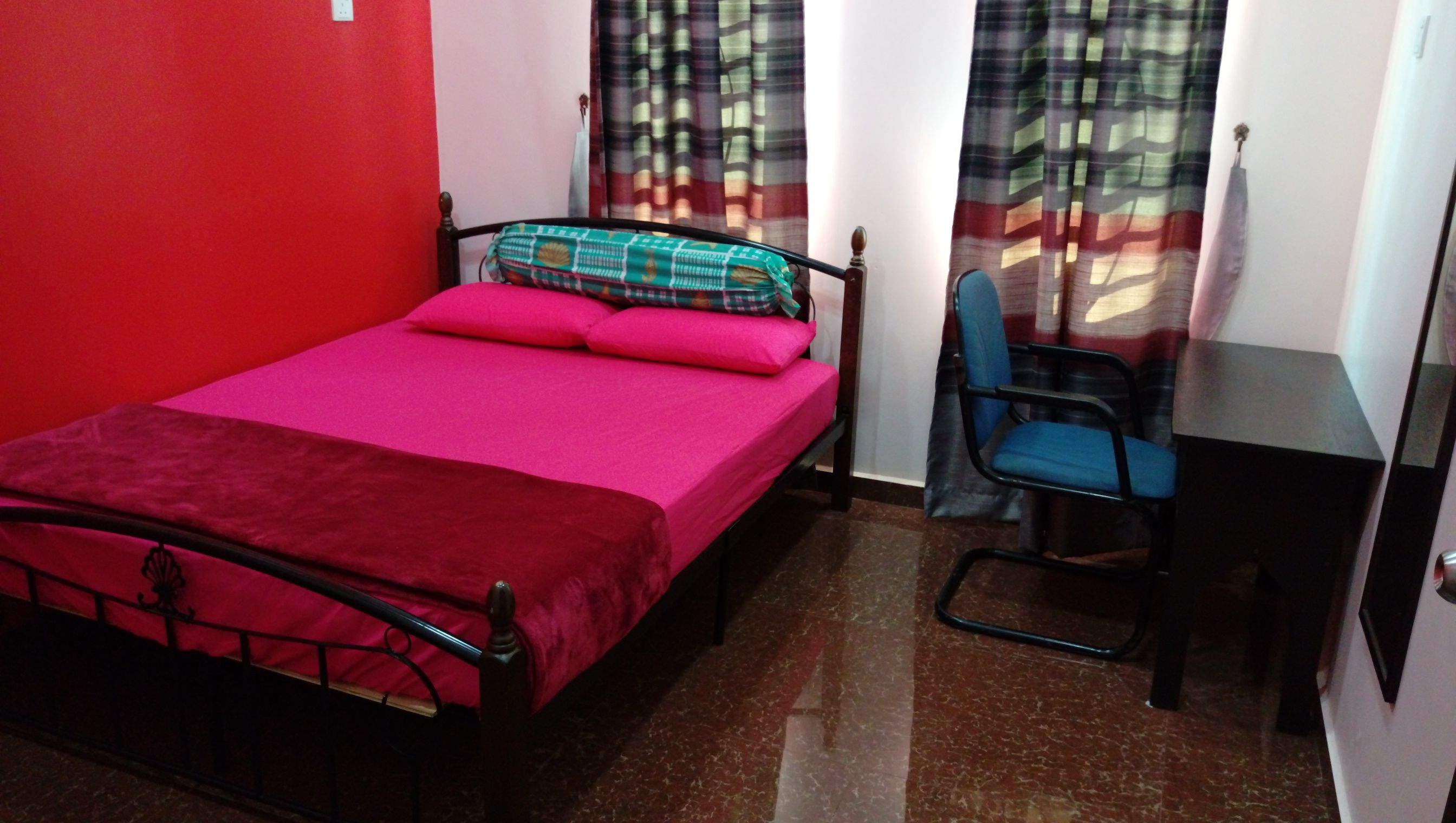 A small, single-bed room with a red wall, a metal bed frame, a blue chair, and a small desk.