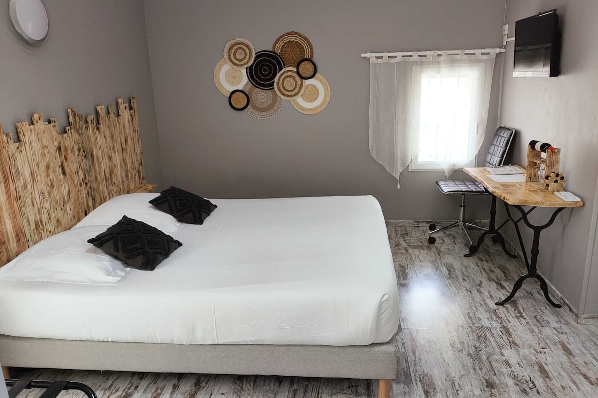 Un dormitorio con cama, cabecera de madera, una obra de arte en la pared, un escritorio con silla, televisor y ventana con cortinas.